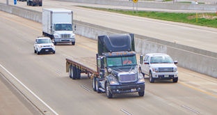 Semi with empty flatbed, four-door pickup truck, box truck and car driving on one side of six-lane divided highway